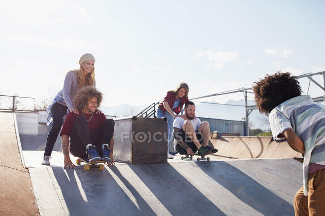 Amigos empujándose unos a otros en patinetas en el soleado parque de skate - foto de stock