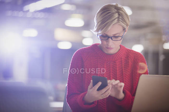 Empresária trabalhando até tarde no laptop, enviando mensagens de texto com telefone celular no escritório escuro — Fotografia de Stock