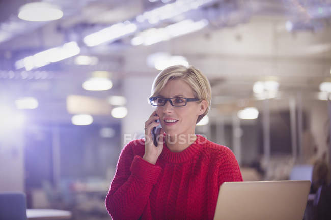 Бізнес-леді розмовляє на мобільному телефоні біля ноутбука в темному офісі, працює пізно — стокове фото