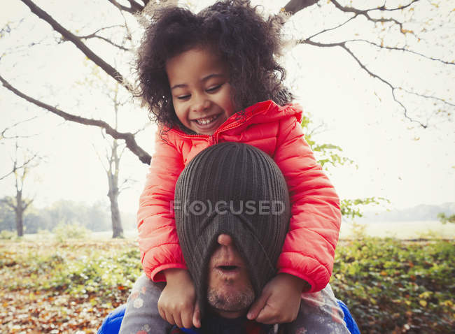 Playful daughter pulling stocking cap over fathers face in autumn park — Stock Photo