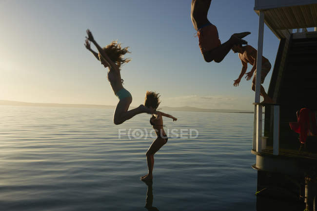 Jóvenes amigos adultos saltando desde la casa flotante de verano en el océano puesta de sol - foto de stock