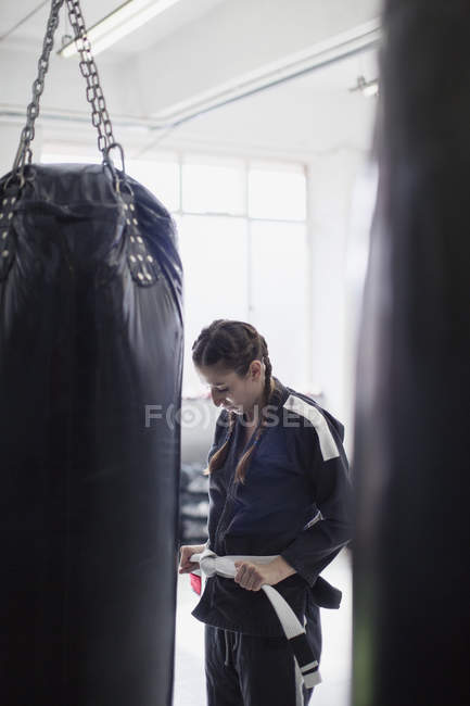 Молодая женщина, завязывающая пояс для дзюдо рядом с боксерской грушей в спортзале — стоковое фото