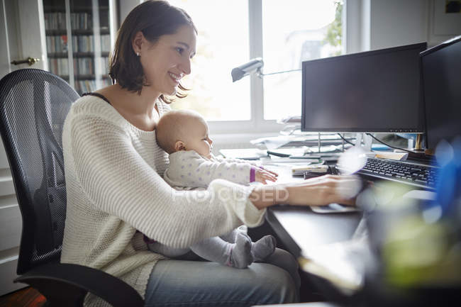 Усміхнена мати тримає доньку, яка працює за столом в домашньому офісі — стокове фото