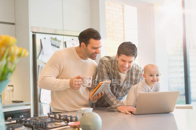 Padres gay masculinos con hijo bebé usando tableta digital y computadora portátil en la cocina - foto de stock