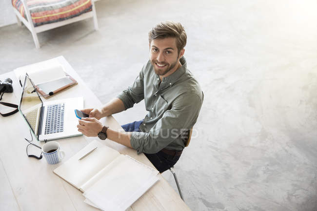 Portrait de jeune homme assis au bureau avec téléphone portable et ordinateur portable — Photo de stock