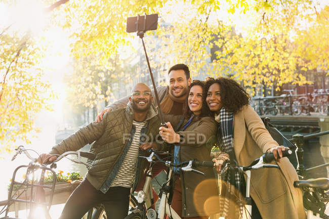 Amigos sorridentes com bicicleta tirando selfie com vara de selfie na ponte urbana — Fotografia de Stock
