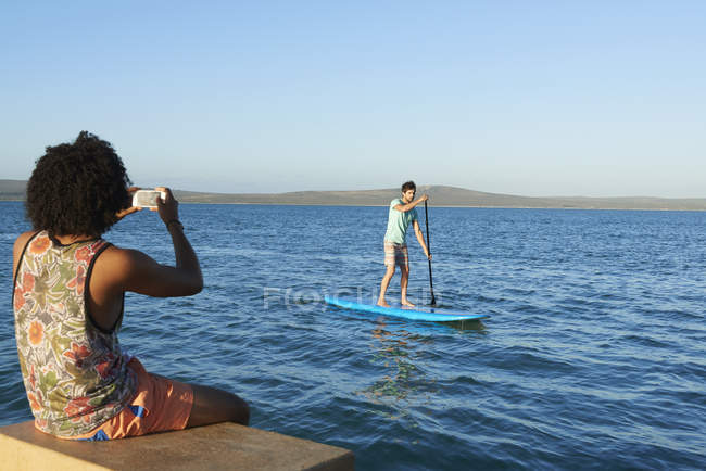 Young man photographing friend paddleboarding on sunny summer ocean — Stock Photo