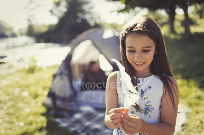 Sorrindo menina segurando penas brancas fora da tenda acampamento ensolarado — Fotografia de Stock