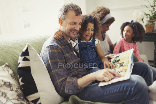Усміхнений багатоетнічний батько читає книгу з дочкою на дивані — стокове фото