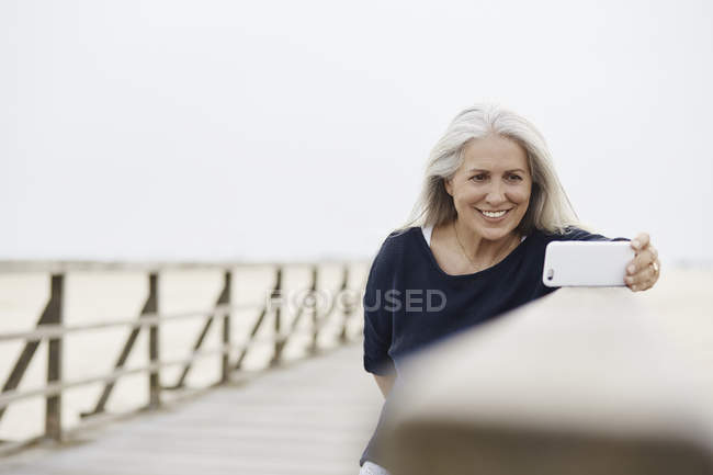 Smiling senior woman taking selfie with camera phone on beach boardwalk — Stock Photo