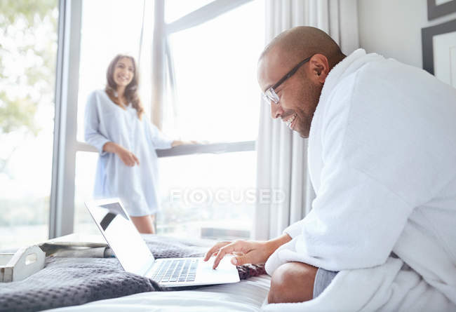 Donna sorridente guardando fidanzato in accappatoio lettura laptop sul letto — Foto stock