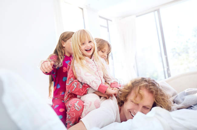 Hijas juguetonas acostadas encima del padre en la cama - foto de stock