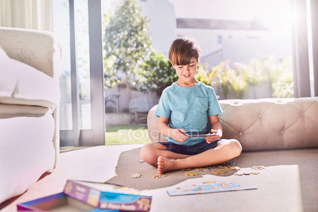 Menino com tablet digital montando quebra-cabeça no chão ensolarado da sala de estar — Fotografia de Stock