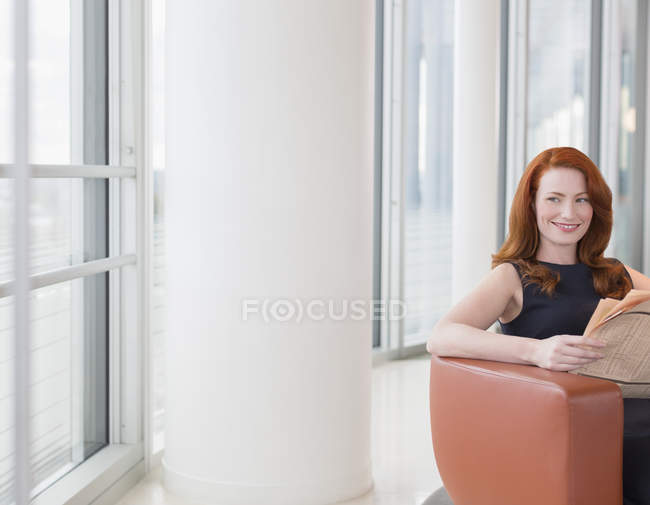 Улыбающаяся деловая женщина с рыжими волосами читает газету в офисе — стоковое фото