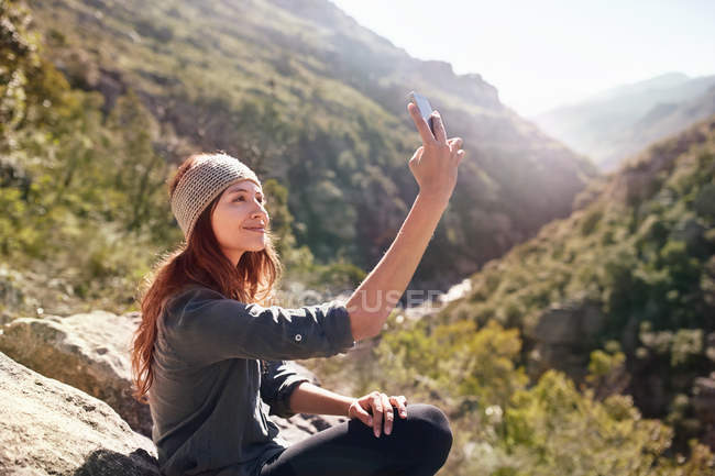Young woman taking selfie with camera phone on sunny, remote rock — Stock Photo