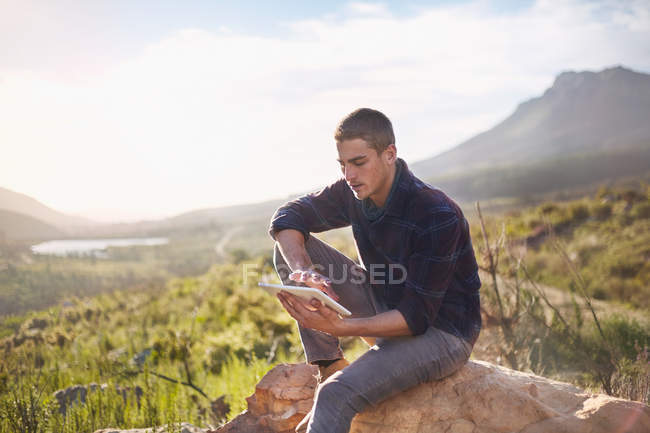 Jeune homme utilisant tablette numérique sur la roche dans la vallée ensoleillée et éloignée — Photo de stock