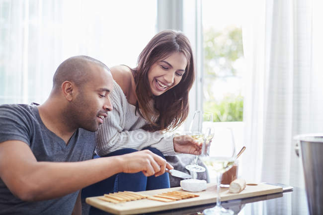 Coppia sorridente che beve vino bianco e mangia cracker e formaggio in soggiorno — Foto stock