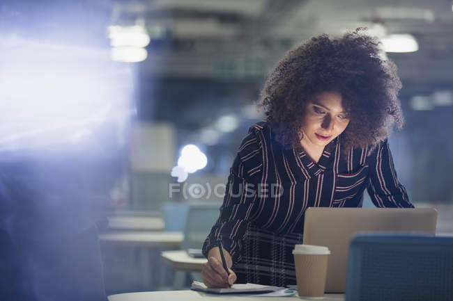 Empresaria enfocada trabajando hasta tarde en el portátil, tomando notas en la oficina oscura - foto de stock