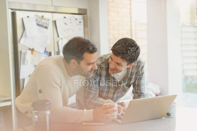 Male gay couple using laptop and drinking coffee in kitchen — Stock Photo