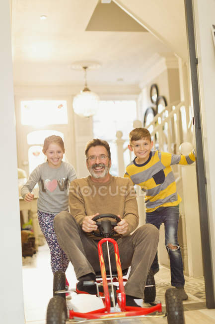 Retrato sorrindo crianças empurrando pai no brinquedo carro no corredor — Fotografia de Stock