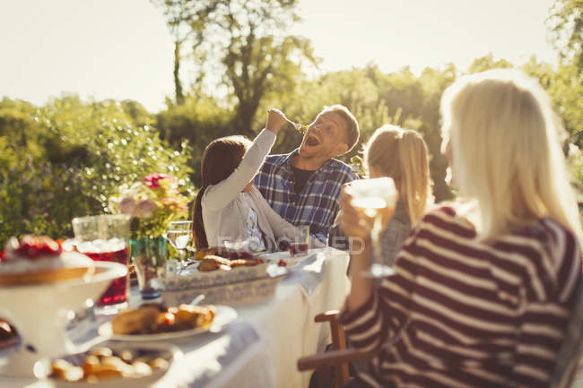Playful daughter feeding father at sunny garden party patio table — Stock Photo