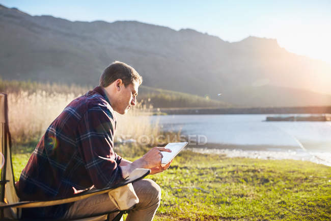 Молодой человек использует цифровые планшеты на солнечном берегу озера — стоковое фото