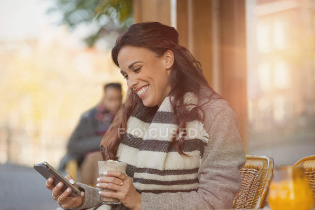 Улыбающаяся женщина пишет смс с мобильного телефона и пьет молочный коктейль в кафе на тротуаре — стоковое фото