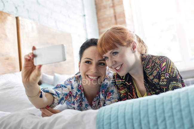 Mulheres sorridentes tomando selfie com telefone câmera deitada na cama — Fotografia de Stock