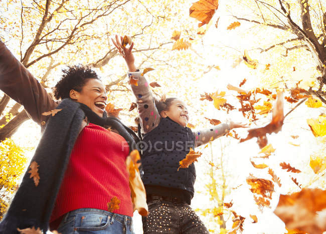 Mère et fille ludiques jetant des feuilles d'automne dans un parc ensoleillé — Photo de stock