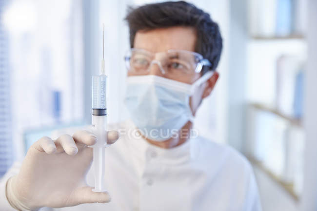 Человек в хирургической маске и халате держит шприц с жидкостью — стоковое фото