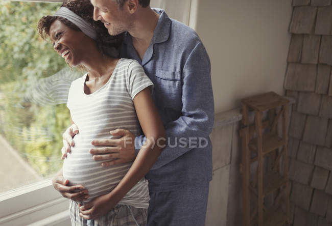 Sonriente pareja embarazada en pijama abrazándose a la ventana - foto de stock