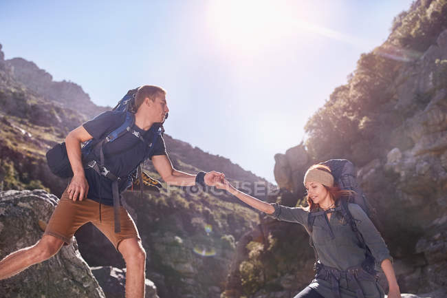 Junger Mann hilft Freundin beim Wandern unter sonnigen, schroffen Klippen — Stockfoto