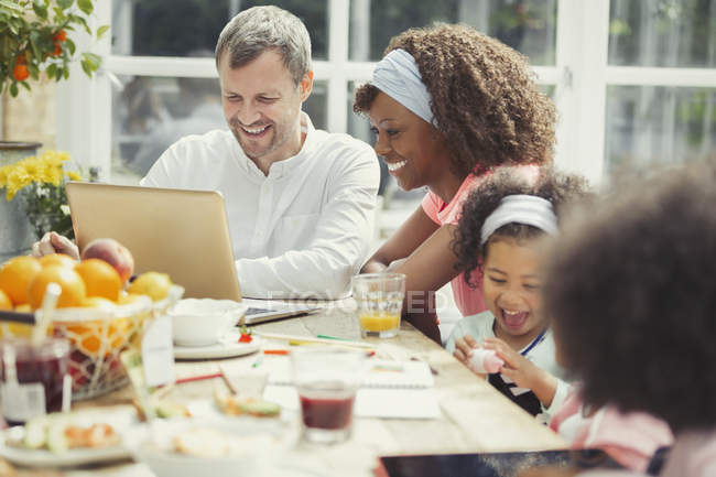 Sonriente joven familia multiétnica utilizando el ordenador portátil y desayunar en la mesa - foto de stock