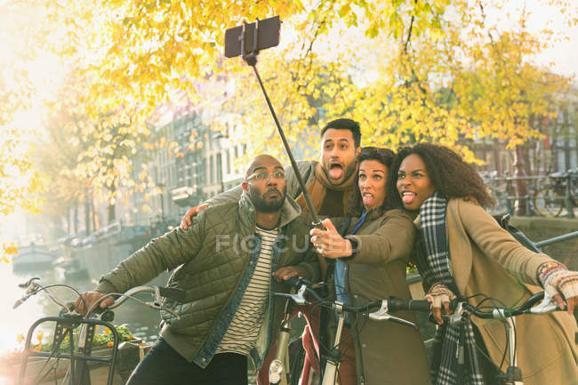 Playful young friends with bicycles making a face taking selfie with selfie stick along autumn canal, Amsterdam — Stock Photo