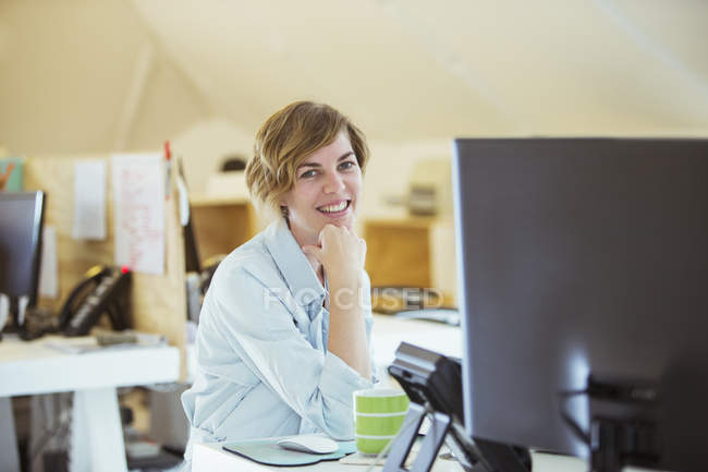 Retrato de la mujer sonriendo en la oficina, sentado en el escritorio con el ordenador - foto de stock