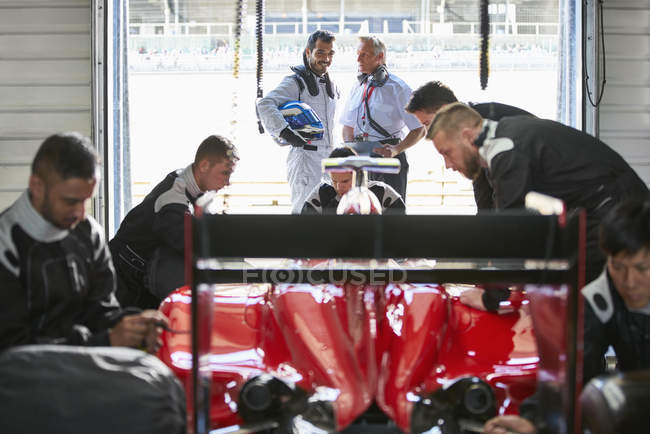 Conductor de Fórmula 1 y gerente hablando detrás de la tripulación de boxes trabajando en el coche de carreras en el garaje de reparación - foto de stock