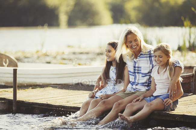 Smiling, affectionate mother and daughters on dock splashing feet in lake — Stock Photo