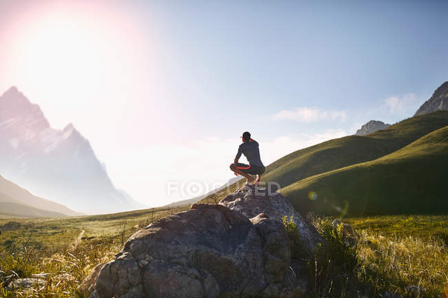 Молодий чоловік вислизає на камінь, дивлячись на сонячний, імітує вид на гори — стокове фото