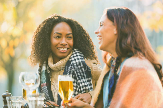 Sonrientes jóvenes amigas bebiendo cerveza en el café de otoño en la acera - foto de stock