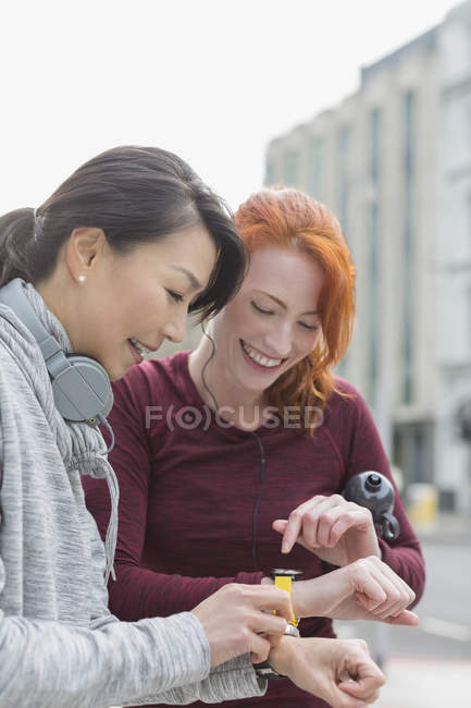 Sorrindo corredores do sexo feminino verificando relógios inteligentes na calçada urbana — Fotografia de Stock