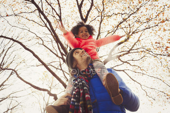 Father carrying daughter on shoulders under tree in autumn park — Stock Photo
