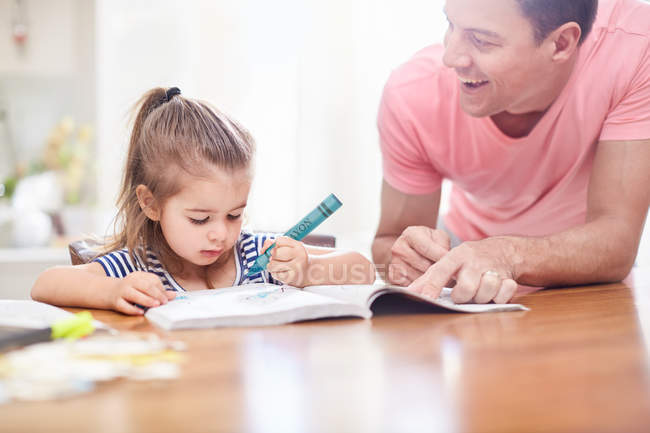 Father watching daughter coloring with crayon and coloring book at table — Stock Photo