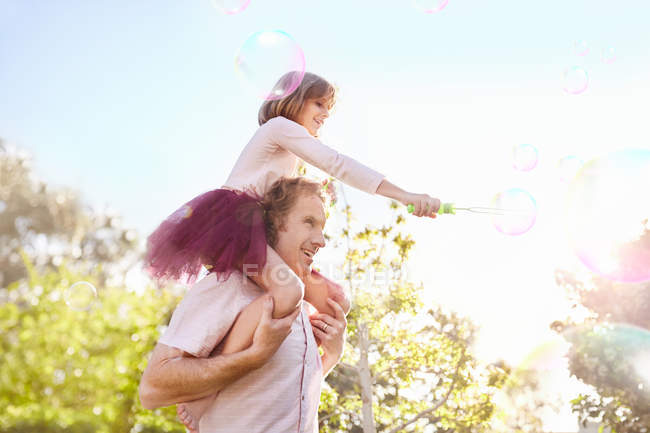 Father carrying daughter with bubble wand on shoulders in sunny summer park — Stock Photo