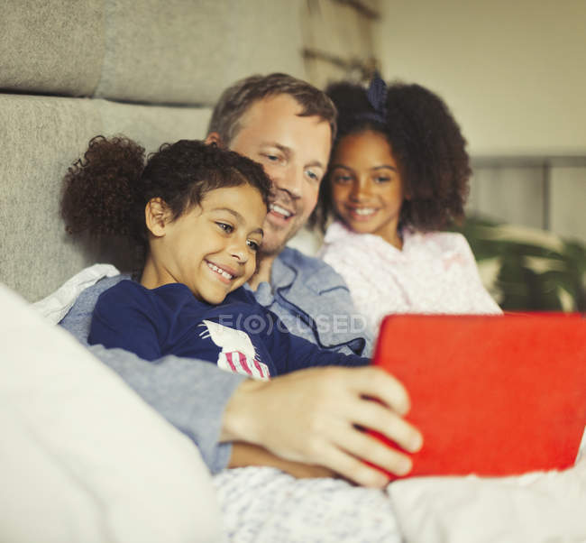 Padre e hijas multiétnicos usando tableta digital en la cama - foto de stock