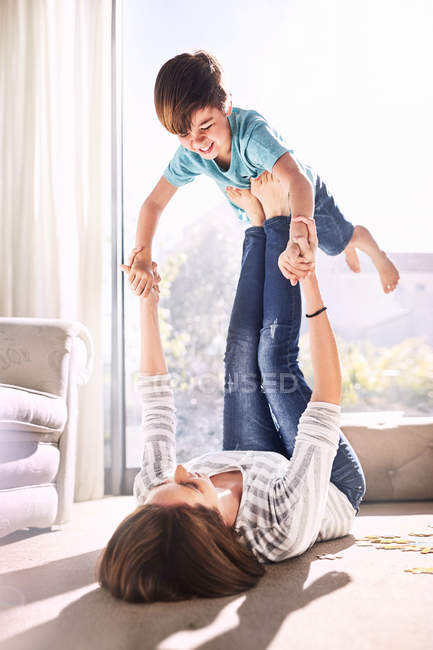 Mãe de levantamento e balanceamento filho em pés no chão da sala de estar ensolarado — Fotografia de Stock