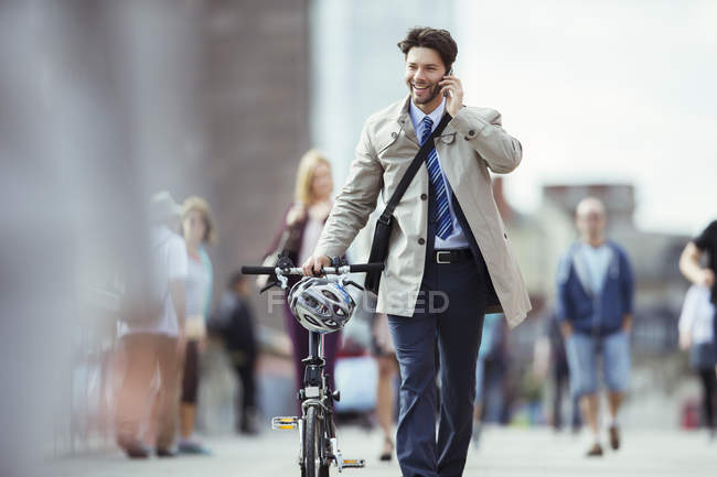 Businessman talking on cell phone pushing bicycle in city — Stock Photo