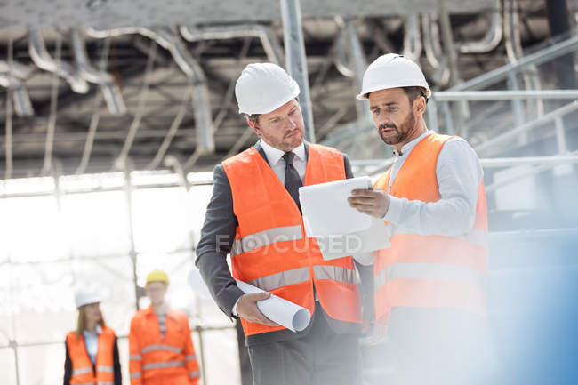 Ingenieros masculinos con planos y portapapeles discutiendo papeleo en el sitio de construcción - foto de stock