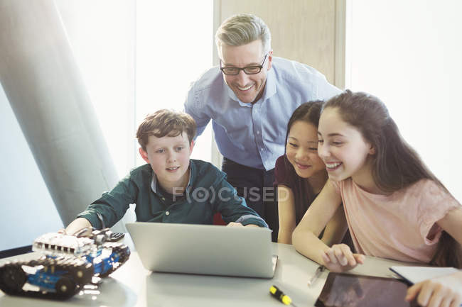 Profesor sonriente y estudiantes programando robótica en la computadora portátil en el aula - foto de stock