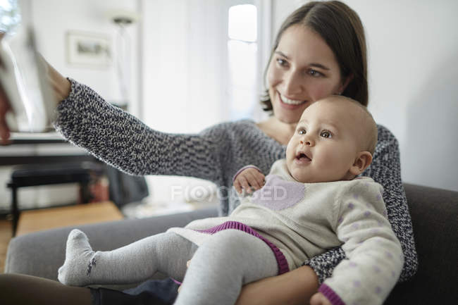 Smiling mother holding baby daughter and taking selfie with camera phone — Stock Photo