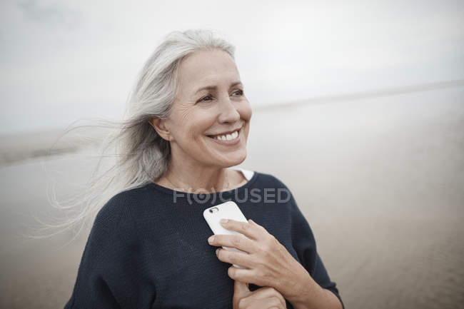 Улыбающаяся пожилая женщина держит мобильный телефон на зимнем пляже — стоковое фото
